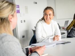 Two students sitting at a desk in a classroom chatting. 