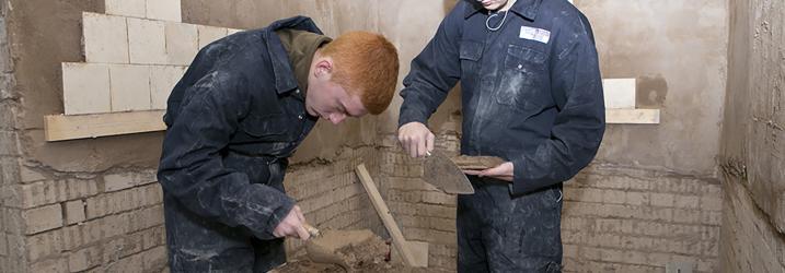 Two students mixing plaster and putting tiles on the wall.