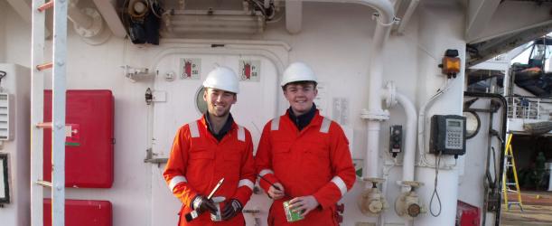 Two engineers in bright orange overalls standing the deck of a ship.