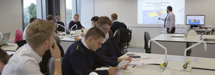 Nautical students in uniform in a classroom with a lecturer standing in front of the class pointing to a board.