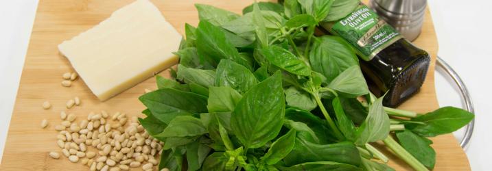 A wooden board with ingredients for pesto on it - parmesan cheese, pine nuts, basil, olive oil and a metal salt grinder.